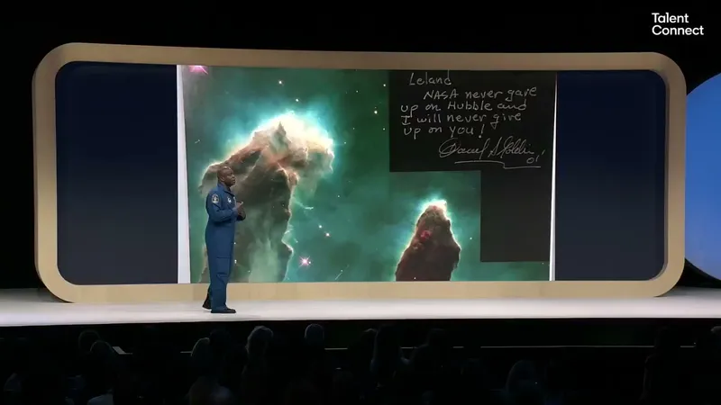 Leland Melvin in a blue flight suit on stage in front of a Hubble image of a nebula with a handwritten dedication from Daniel Goldin