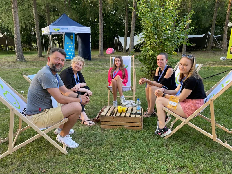 Five people sitting on deckchairs around a wooden crate on the grass among trees, with Campus Polska Przyszłości tents in the background
