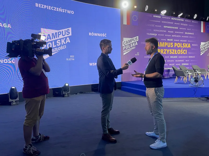 Rafał Szymański giving a TV interview in front of the stage at Campus Polska Przyszłości 2022 in Olsztyn: a reporter holds a microphone, a camera operator stands beside them, the panelists' armchairs are in the background