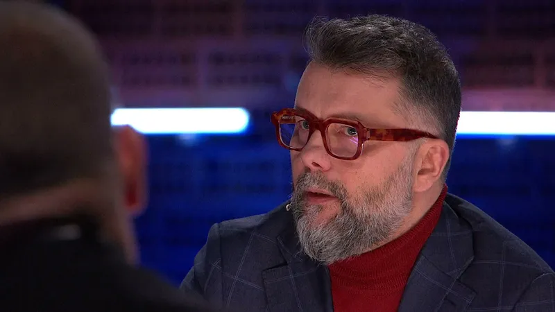 Rafał Szymański w czerwonych okularach i granatowej marynarce mówi w studiu programu Hala odlotów w TVP