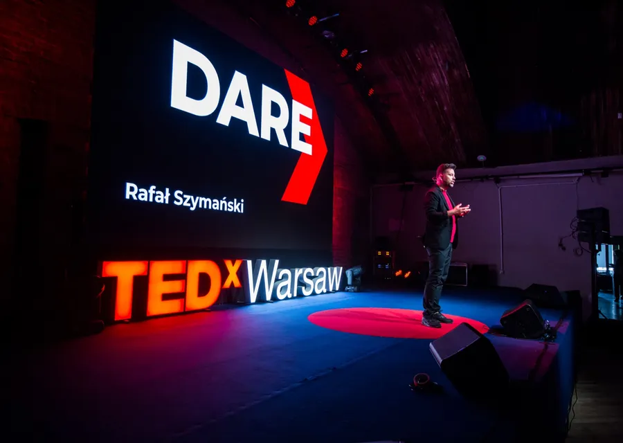 Rafał Szymański on the TEDxWarsaw stage next to a screen with the word DARE and his name